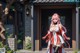 A woman in a red and white outfit stands outside a house with a stone wall and greenery.